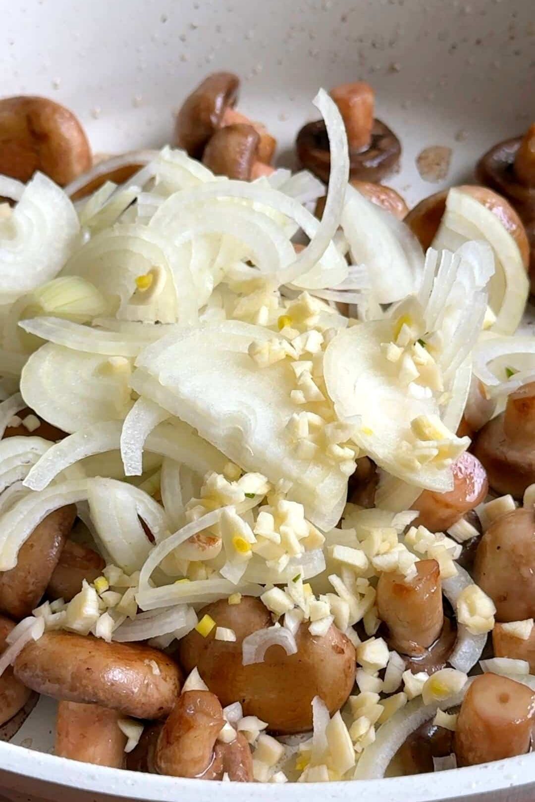 Browned mushrooms in a skillet with freshly added sliced onions and chopped garlic, cooking until the onions turn translucent.
